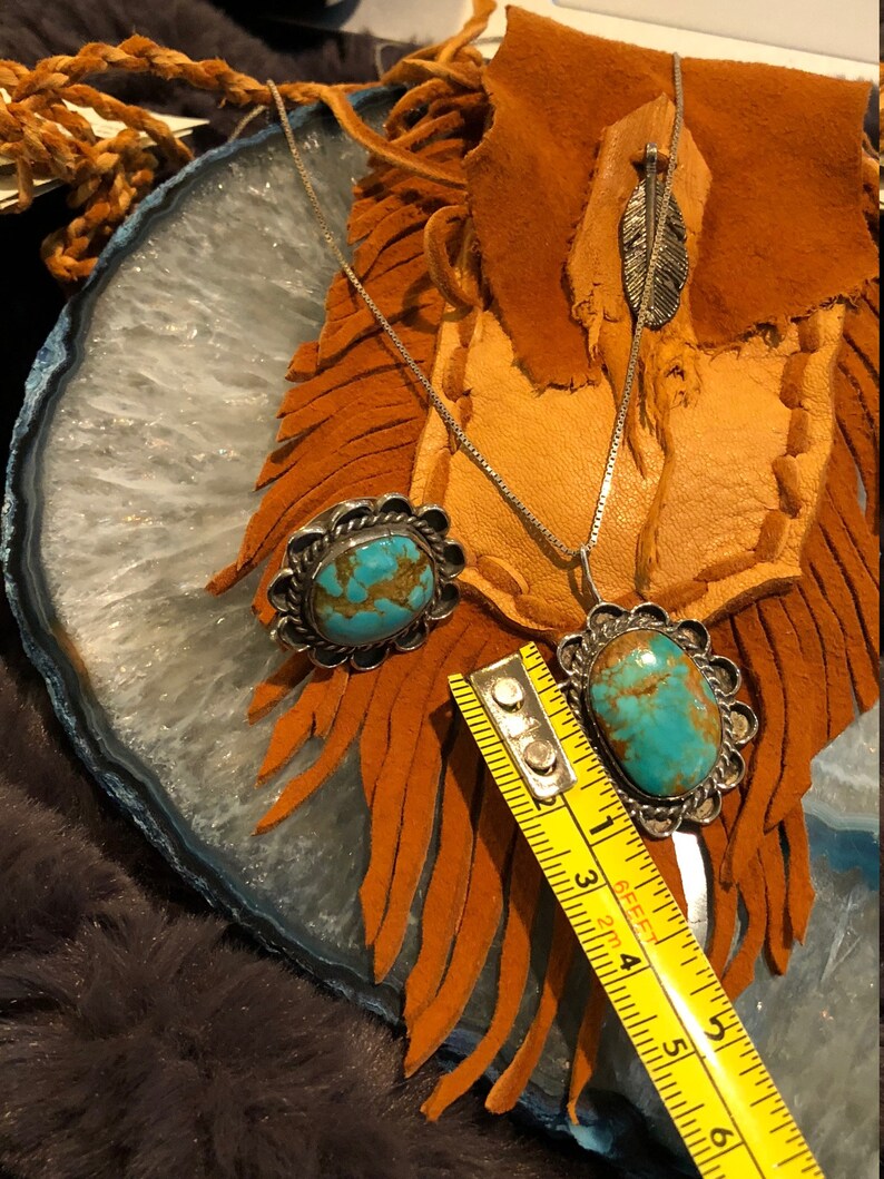 May include: A silver necklace with a turquoise stone pendant and a matching turquoise stone ring. The necklace is on a brown leather background with fringe.