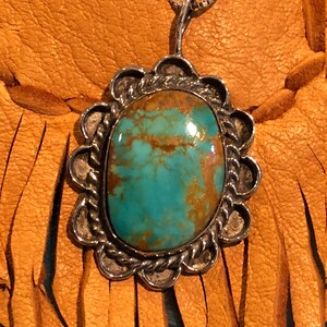 May include: A silver pendant featuring a turquoise stone set within a scalloped border. The pendant hangs from a silver chain and is shown against a brown leather backdrop.