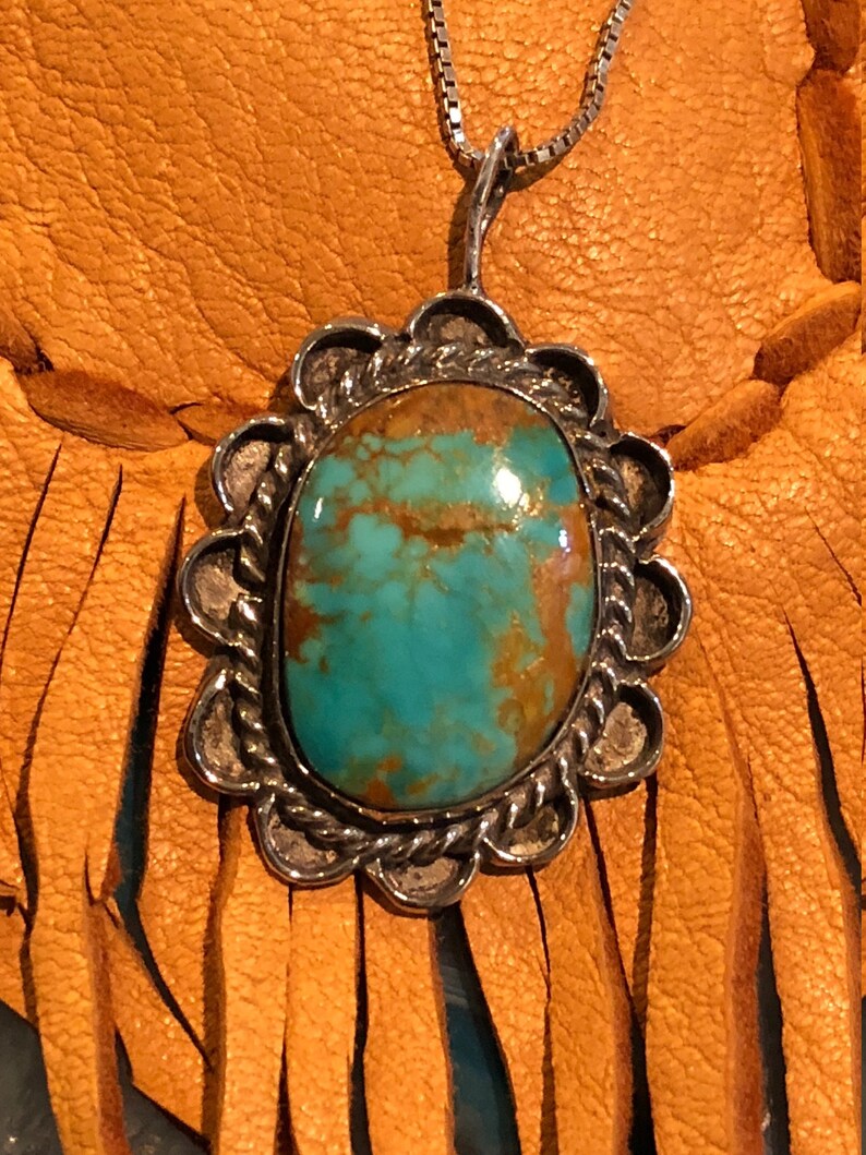 May include: A silver pendant featuring a turquoise stone set within a scalloped border. The pendant hangs from a silver chain and is shown against a brown leather backdrop.