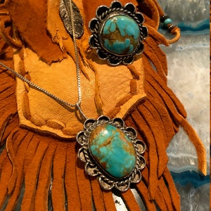 May include: A turquoise pendant necklace and ring set with silver settings. The turquoise stones are a vibrant blue-green colour with brown and gold inclusions. The necklace is on a silver chain and the ring is set in a silver band with a decorative border.