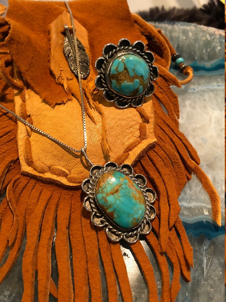 May include: A turquoise pendant necklace and ring set with silver settings. The turquoise stones are a vibrant blue-green colour with brown and gold inclusions. The necklace is on a silver chain and the ring is set in a silver band with a decorative border.