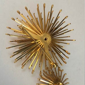 May include: A pair of gold-toned brooches in the shape of a starburst. Each brooch has a pin back and a cluster of thin, pointed metal rays.