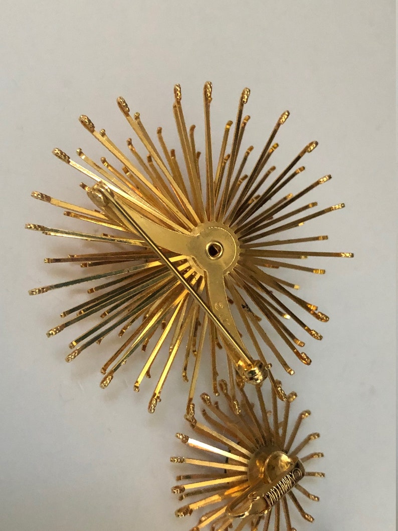 May include: A pair of gold-toned brooches in the shape of a starburst. Each brooch has a pin back and a cluster of thin, pointed metal rays.