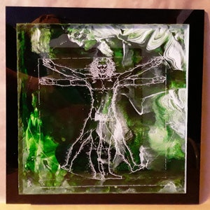 May include: A white outline of the Vitruvian Man, a famous drawing by Leonardo da Vinci, is etched into a green and white resin art piece. The resin is encased in a black frame.