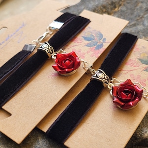 May include: Two black velvet choker necklaces with silver rose charms. The roses are red and have a detailed design.