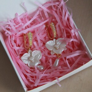 May include: A pair of gold hoop earrings with white orchid flower charms. The earrings are displayed in a pink tissue paper-filled box.