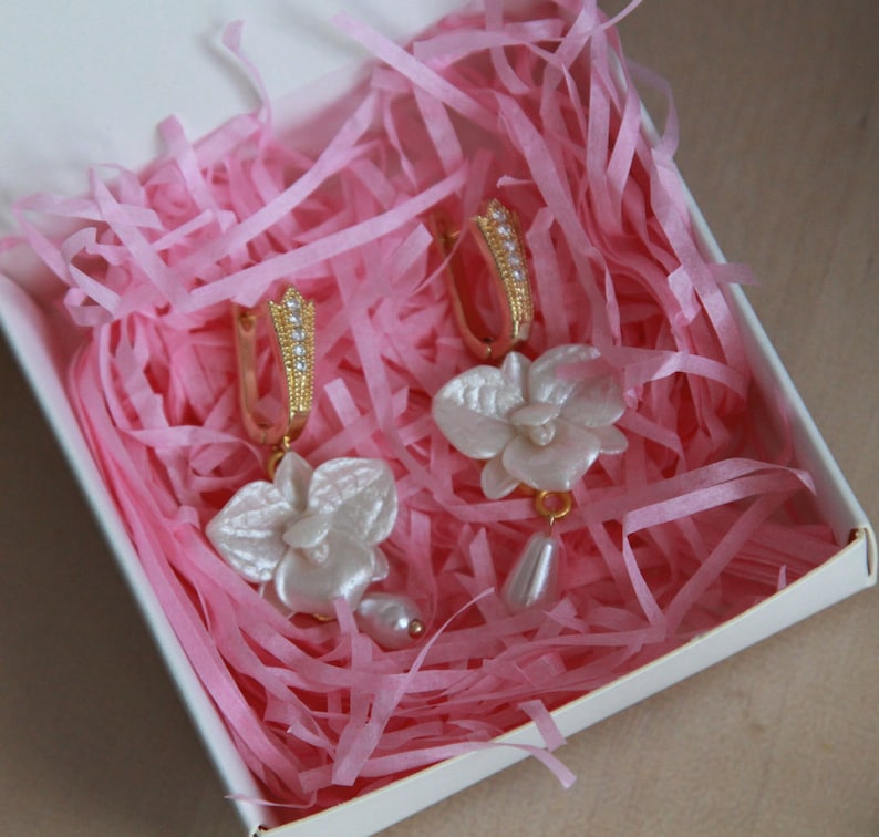 May include: A pair of gold hoop earrings with white orchid flower charms. The earrings are displayed in a pink tissue paper-filled box.