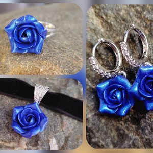 May include: A jewelry set featuring a ring, a pendant on a black velvet ribbon, and a pair of hoop earrings. Each piece displays a metallic blue rose with layered petals. The silver-toned hoop earrings are accented with small clear stones.