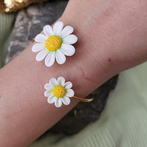 May include: A gold-coloured bracelet with two daisy flower features. The daisies have white petals and yellow centres. The bracelet wraps around the wrist.
