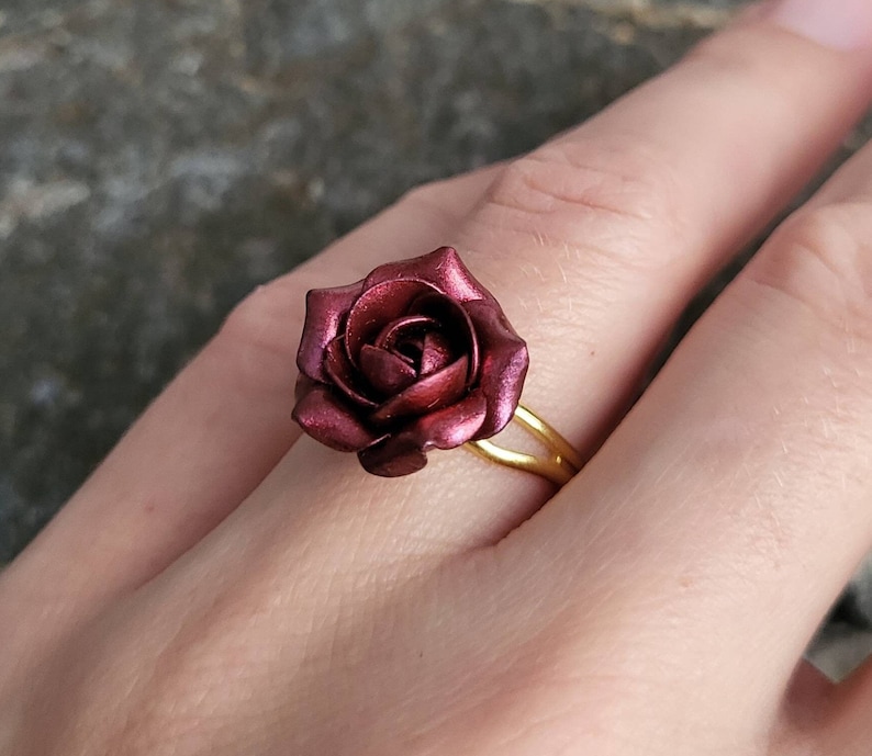 May include: A gold ring with a burgundy rose-shaped design. The rose is made of polymer clay and has a metallic finish.