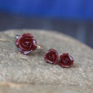 May include: A jewelry set featuring a ring and two stud earrings, all with a burgundy rose design. The ring has a silver band. The roses have a subtle iridescent sheen. The set is displayed on a gray stone surface.