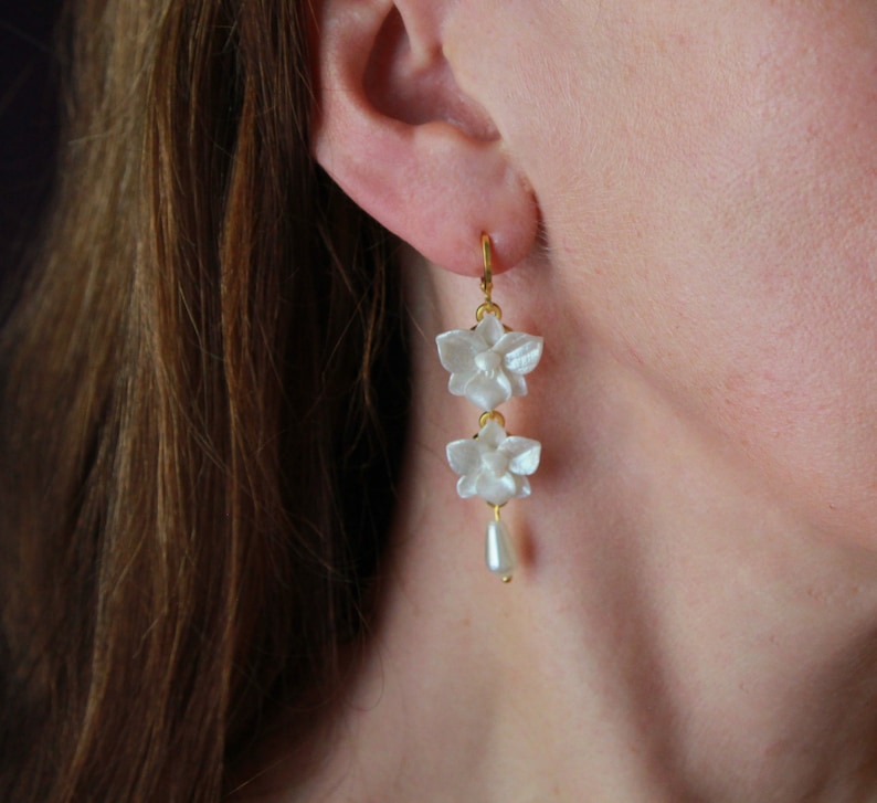 May include: White flower dangle earrings with a pearl drop. The earrings are made of gold metal and have a delicate design.