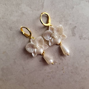May include: A pair of gold earrings with white orchid flower charms and pearl drop accents.