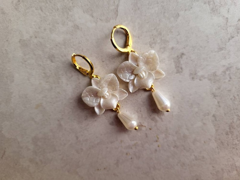 May include: A pair of gold earrings with white orchid flower charms and pearl drop accents.
