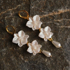 May include: A pair of gold earrings with three white flower-shaped charms and a pearl drop on each earring.