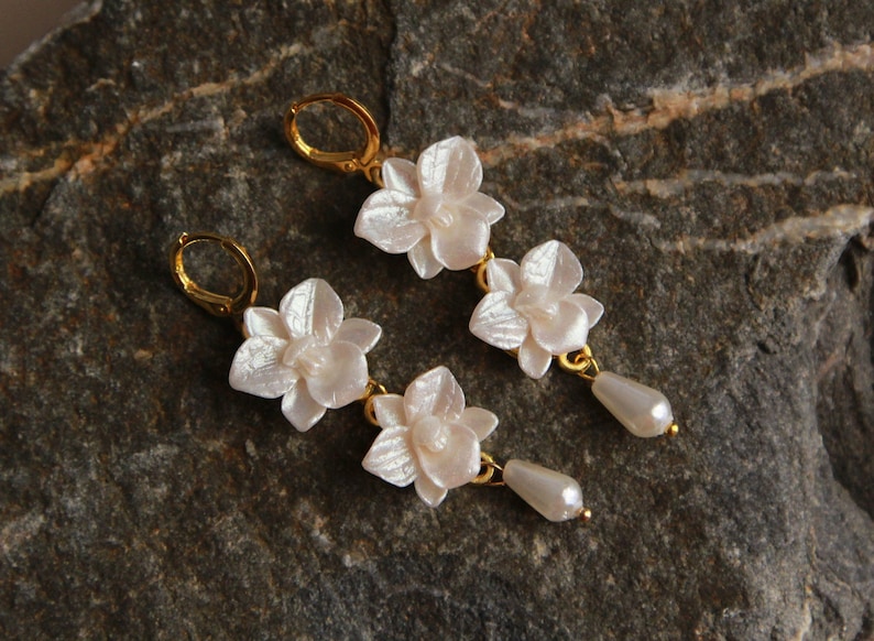 May include: A pair of gold earrings with three white flower-shaped charms and a pearl drop on each earring.
