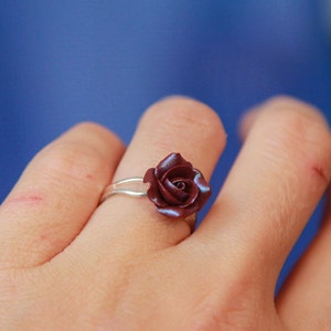 May include: A silver ring featuring a burgundy rose. The rose has detailed petals and a subtle iridescent sheen. The ring is worn on a finger against a blue background, showcasing the jewelry's design and craftsmanship.
