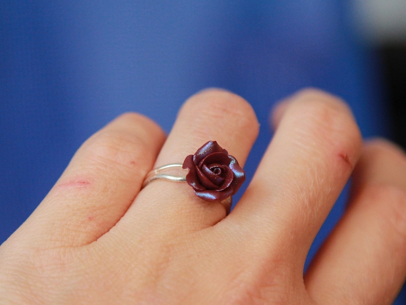 May include: A silver ring featuring a burgundy rose. The rose has detailed petals and a subtle iridescent sheen. The ring is worn on a finger against a blue background, showcasing the jewelry's design and craftsmanship.