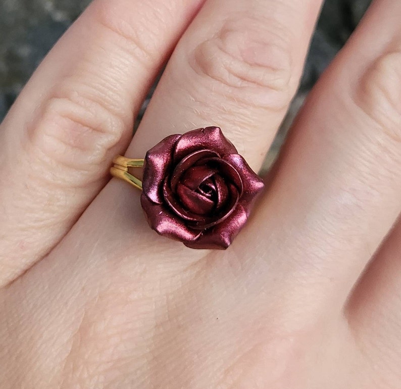 May include: A gold ring with a burgundy rose-shaped design. The rose is made of polymer clay and has a metallic finish.