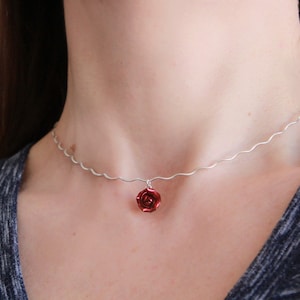 Könnte beinhalten: Eine silberne Halskette mit einer wellenförmigen Kette und einem kleinen, roten Rosenanhänger. Die Rose hat detaillierte Blütenblätter und hängt an der Kette. Die Halskette wird um den Hals getragen.