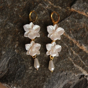 May include: A pair of gold earrings with three white orchid flower charms and a pearl drop on each earring.