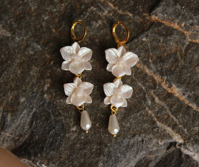 May include: A pair of gold earrings with three white orchid flower charms and a pearl drop on each earring.