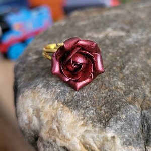 May include: A gold ring with a deep red rose attached to the band. The rose is made of metal and has a shiny finish.