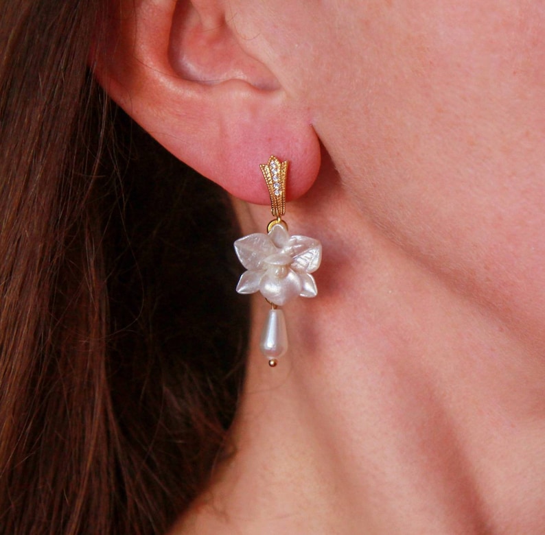 May include: Gold earrings with a white flower design and a pearl drop. The flower is made of a pearlescent material and has a delicate, feminine look.