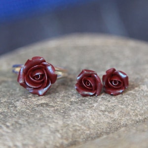 May include: A jewelry set featuring a burgundy rose ring and matching stud earrings. The ring has a silver band. The rose petals have a subtle iridescent sheen. The earrings are small, detailed rose buds. The set is displayed on a textured, gray surface.