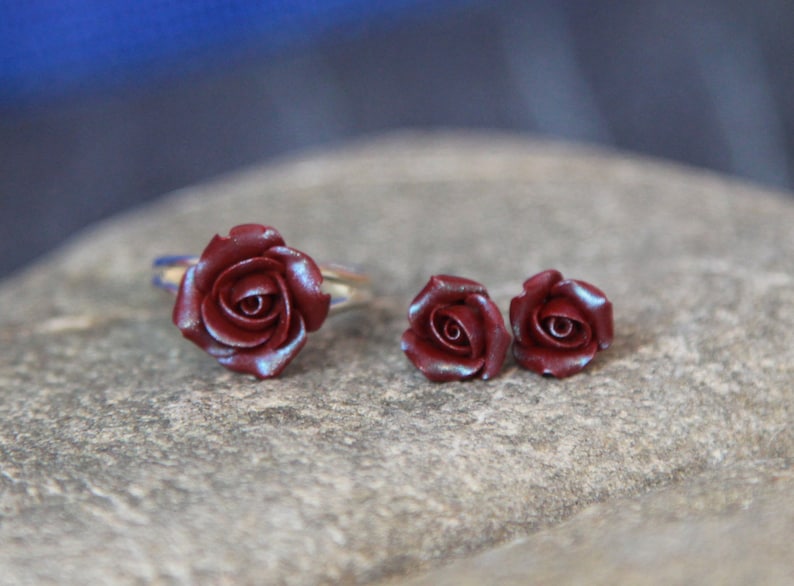 May include: A jewelry set featuring a burgundy rose ring and matching stud earrings. The ring has a silver band. The rose petals have a subtle iridescent sheen. The earrings are small, detailed rose buds. The set is displayed on a textured, gray surface.