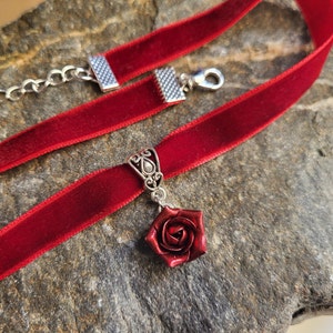 Può includere: Un girocollo in velluto rosso con un ciondolo a forma di rosa argentata. La rosa è dettagliata e ha un aspetto vintage.
