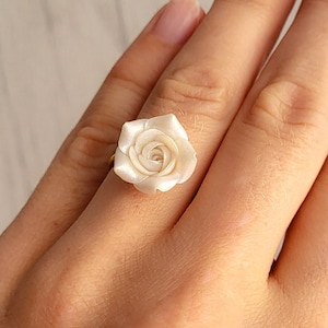 Puede incluir: Un anillo de rosa blanca con una banda dorada. La rosa está hecha de un material nacarado y tiene un diseño delicado y femenino.