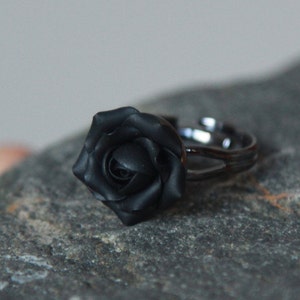 May include: A black rose ring. The ring features a detailed, three-dimensional rose design in matte black. The adjustable band is also black, creating a cohesive and elegant look. The ring is set against a neutral background.