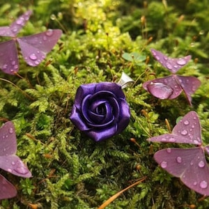 Könnte beinhalten: Nahaufnahme eines lila Rosenrings vor dem Hintergrund von grünem Moos und lila Kleeblättern. Der Ring zeigt ein detailliertes, stoffähnliches Rosendesign. Wassertropfen sind auf den Blättern sichtbar.