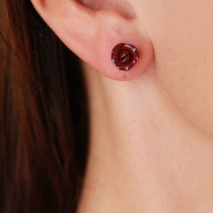 May include: A single dark red rose stud earring. The earring is a miniature, three-dimensional rose shape, with detailed petals. The earring is worn in an ear piercing. The background is a blurred skin tone.