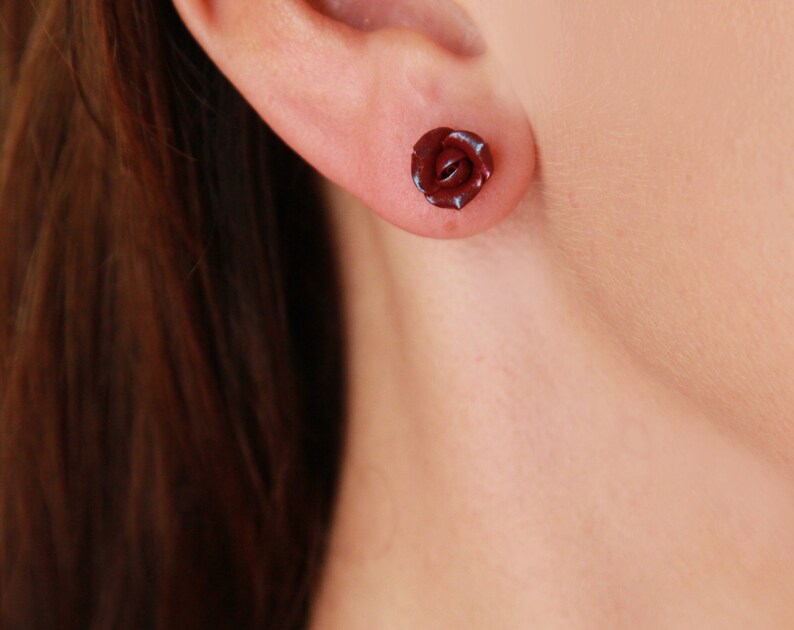 May include: A single dark red rose stud earring. The earring is a miniature, three-dimensional rose shape, with detailed petals. The earring is worn in an ear piercing. The background is a blurred skin tone.