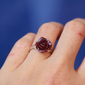 May include: A silver ring featuring a dark red rose. The rose is the focal point, with detailed petals. The ring is set against a blue background, highlighting the jewelry's design. A close-up shot emphasizes the ring's craftsmanship.