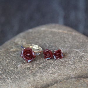 May include: A jewelry set featuring a ring and matching stud earrings. The ring and earrings have a silver-colored band and are adorned with dark red rose-shaped embellishments. The set is displayed on a gray stone surface.