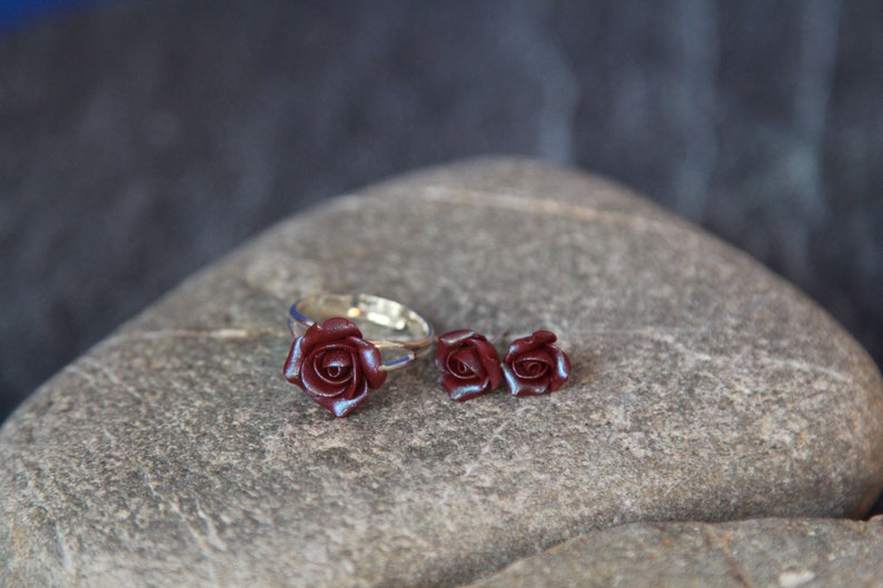 May include: A jewelry set featuring a ring and matching stud earrings. The ring and earrings have a silver-colored band and are adorned with dark red rose-shaped embellishments. The set is displayed on a gray stone surface.
