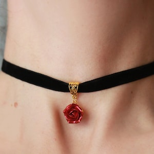 Può includere: Un girocollo in velluto nero con un ciondolo a forma di rosa dorata. La rosa è rossa e presenta un design dettagliato.