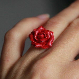 Può includere: Un anello a forma di rosa rossa in argilla polimerica. La rosa è dettagliata con linee nere e ha un aspetto realistico.