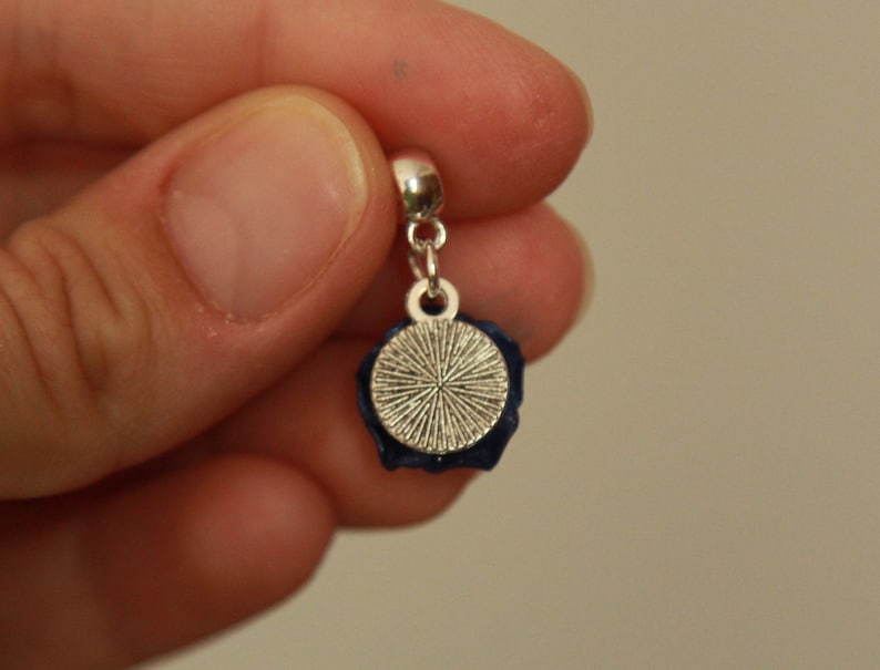 May include: A silver charm with a blue flower design and a round, silver, textured disc. The charm is attached to a silver chain.
