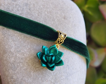 May include: A dark green velvet choker necklace with a gold-colored clasp and a teal succulent pendant. The choker is displayed on a light gray surface, with green foliage in the background. The succulent pendant is a detailed, three-dimensional design.