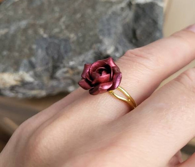 May include: A gold ring with a burgundy rose-shaped design. The ring is worn on a person's finger.