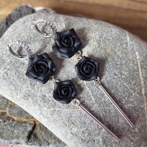 Könnte beinhalten: Ohrringe mit schwarzen Rosenanhängern und silberfarbenen Akzenten. Die Ohrringe haben ein Creolen-Design mit kleinen, klaren Steinen. Die Rosen sind ein dunkles, mattes Schwarz, und die Ohrringe werden auf einer grauen Steinoberfläche präsentiert.