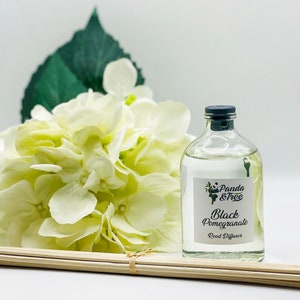 Puede incluir: Una botella de vidrio transparente con una tapa negra y una etiqueta blanca que dice "Panda & Tree Black Pomegranate Reed Diffuser". La botella está sobre una superficie blanca con un ramo de flores blancas y palitos de madera.