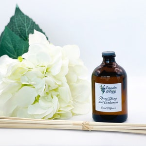 Op de afbeelding: Een bruine glazen fles met een zwart etiket waarop "Panda & Tree Ylang Ylang and Cardamom Reed Diffuser" staat. De fles staat op een wit oppervlak met een boeket witte bloemen en een set houten diffuserstokjes.