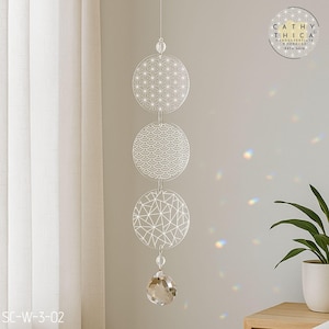 Könnte beinhalten: Dekorative Hängeornament mit drei kreisförmigen, gemusterten Scheiben, die an einer klaren Perle aufgehängt sind. Die obere Scheibe hat ein Flower-of-Life-Design, die mittlere Scheibe ein Wellenmuster und die untere Scheibe ein geometrisches Design. Am unteren Ende hängt ein Kristallprisma.