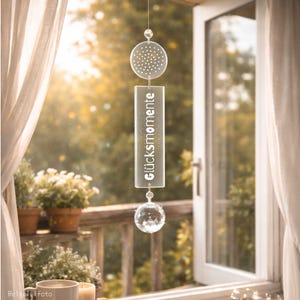 Könnte beinhalten: Ein klarer Windspiel mit dem deutschen Text "Glücksmomente" (Glücksmomente). Es hat eine kreisförmige Oberseite mit kleinen Löchern, einen rechteckigen Mittelteil und einen Kristallkugel am Boden. Der Hintergrund zeigt ein Fenster mit Blick auf einen sonnigen Garten.
