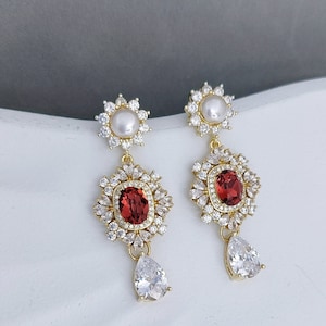 May include: A pair of gold earrings with a red oval gemstone surrounded by clear rhinestones, a white pearl, and a teardrop-shaped clear rhinestone.